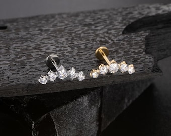 May include: Two ear piercing studs. One stud is silver-toned, the other is gold-toned. Each stud features a row of clear, sparkling gemstones. The studs are displayed on a dark, textured surface.