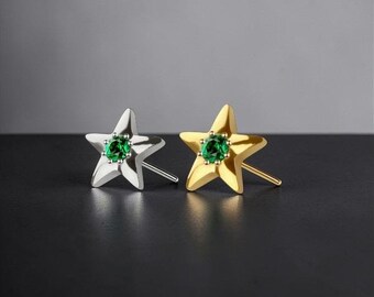 May include: Two star-shaped stud earrings, one silver and one gold, each featuring a vibrant green gemstone at the center. The earrings are displayed on a dark surface, showcasing their metallic shine and gemstone sparkle. Jewelry.
