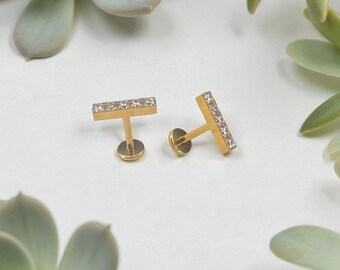 May include: A pair of gold-colored bar stud earrings, each adorned with a row of small, clear crystals. The earrings are displayed on a white surface, surrounded by green succulent plants.