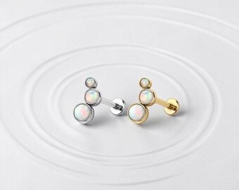 May include: Two pieces of jewelry, one silver and one gold, each featuring three opal-like stones. The stones are arranged in a cluster, with the largest at the base and the smallest at the top. The jewelry is designed for ear piercings.