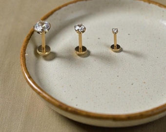 May include: Three gold-colored lip piercing studs with clear, round gemstones are displayed on a speckled, off-white plate with a brown rim. The studs vary in size, showcasing a minimalist jewelry design.