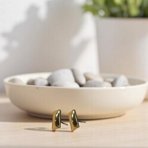 May include: A pair of gold-colored stud earrings with a geometric design. The earrings are rectangular with a slightly angled top. They are displayed on a light-colored wooden surface, with a shallow bowl of stones in the background.
