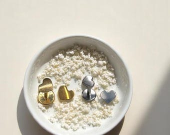 May include: A white bowl filled with small, white, textured objects, containing four heart-shaped jewelry pieces. Two are gold-toned, and two are silver-toned. The earrings are in various styles, including studs and a post-back design.