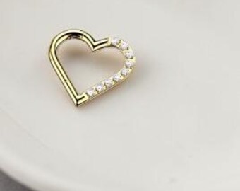 May include: A gold-colored heart-shaped earring or charm, with a row of small, clear gemstones along one side. The jewelry piece is set on a white, heart-shaped dish, against a soft gray background.