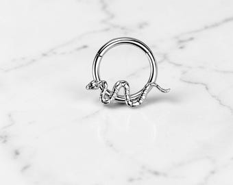 May include: A silver-colored hinged hoop earring with a snake design. The snake is coiled around the bottom of the hoop, with its head and tail extending slightly. The earring is made of a shiny metal.