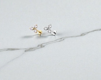 May include: Two small stud earrings with a floral design, each featuring three clear, sparkling gemstones. One earring is gold-toned, the other silver-toned. The earrings are displayed on a white and gray marble surface.