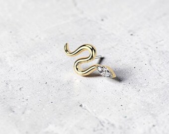 May include: A gold-colored snake-shaped earring with a clear, marquise-cut gemstone at the end. The earring is set against a light gray, textured background. The snake design is detailed and elegant.