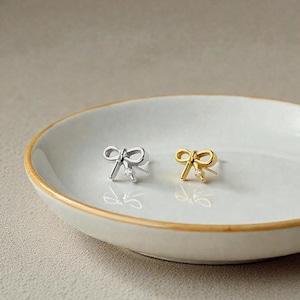 May include: A pair of bow-shaped stud earrings, one silver and one gold, each with a small, clear gemstone at the center. The earrings are displayed on a white, oval-shaped dish with a gold rim.