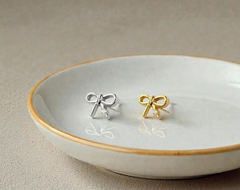 May include: A pair of bow-shaped stud earrings, one silver and one gold, each with a small, clear gemstone at the center. The earrings are displayed on a white, oval-shaped dish with a gold rim.