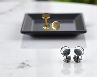 May include: Two pairs of earrings. One pair is gold-colored, with a flat, round stud and a post. The other pair is silver-colored, with a textured, domed stud and a post. The earrings are displayed on a black tray.