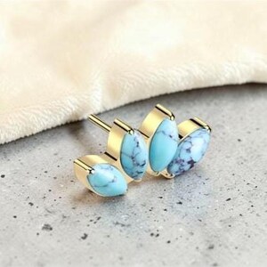 May include: Gold-toned stud earrings featuring oval turquoise stones with dark blue veining. The earrings have a post back and are set against a neutral background. The earrings are a jewelry item.