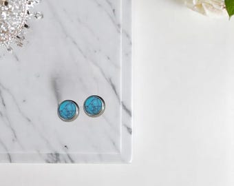 May include: A pair of round stud earrings featuring a bright turquoise stone with dark grey veining set in a polished silver-toned metal frame. The earrings are circular in shape and display a smooth, flat surface with a distinct marbled pattern.