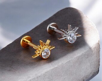 May include: Two flat-back earrings with a sunburst design. One earring is gold-toned, the other is silver-toned. Each features a clear, round gemstone at the center of the sunburst. The earrings are displayed on a gray, textured surface.
