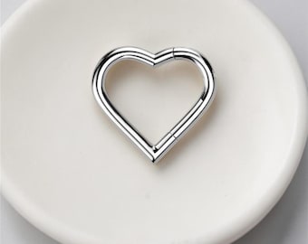 May include: A shiny, silver-colored heart-shaped clasp. The clasp is made of metal and has a hinged opening. The heart is resting on a white, slightly textured surface.