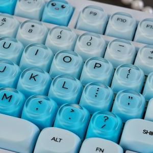 Puede incluir: Primer plano de un teclado con teclas azul claro y blanco. Las teclas tienen tapas transparentes con letras, números y símbolos. Algunas teclas tienen caras de dibujos animados lindos. El teclado es de color azul claro.