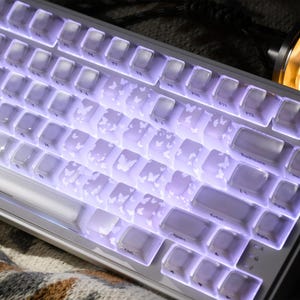 Peut inclure: Un clavier mécanique blanc avec des touches ornées de motifs de papillons. Les touches sont rétroéclairées d'une douce lueur violette. Le clavier a une disposition standard avec des touches de fonction et un pavé numérique.