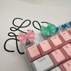May include: A close-up of a mechanical keyboard with pink keycaps and two bow-shaped keycaps. One bow is pink, the other is green. The keyboard has white and gray keys, with a white background and a black logo.