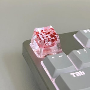 May include: A custom keyboard keycap with a clear resin design. The keycap is filled with red dried flowers. The keycap is on a gray keyboard with white lettering.