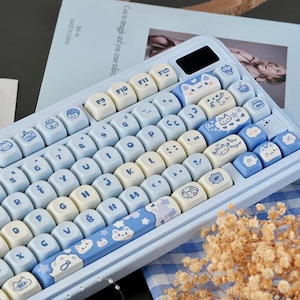 May include: A light blue mechanical keyboard with white and blue keycaps. The keycaps feature cat and cloud designs. The keyboard has a small black screen and is on a light blue surface. The keyboard is surrounded by a blue and white checkered cloth and dried flowers.