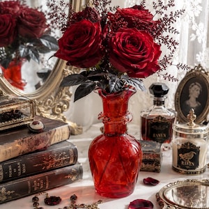 May include: A vibrant red glass vase holds a bouquet of deep red roses and dark foliage. The vase sits on a marble surface surrounded by antique books, jewelry, and decorative items, creating a gothic aesthetic.