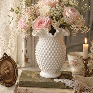 May include: A white hobnail vase filled with pink roses and delicate white flowers. The vase sits atop a stack of vintage books, with a framed picture and a lit candle in the background. A romantic, floral arrangement.