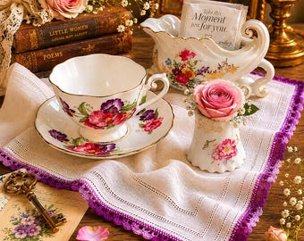 May include: A vintage tea set with a floral design, including a teacup, saucer, and creamer. A small vase holds a pink rose. The scene is set on a white cloth with a purple lace trim, with antique books and a candle in the background.