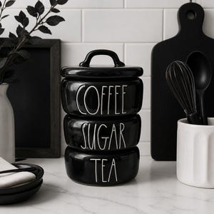 May include: Black ceramic kitchen canisters stacked with the words "COFFEE", "SUGAR", and "TEA" in white. A black handle is on top. Other kitchen items include a cutting board, utensils, and a white vase.