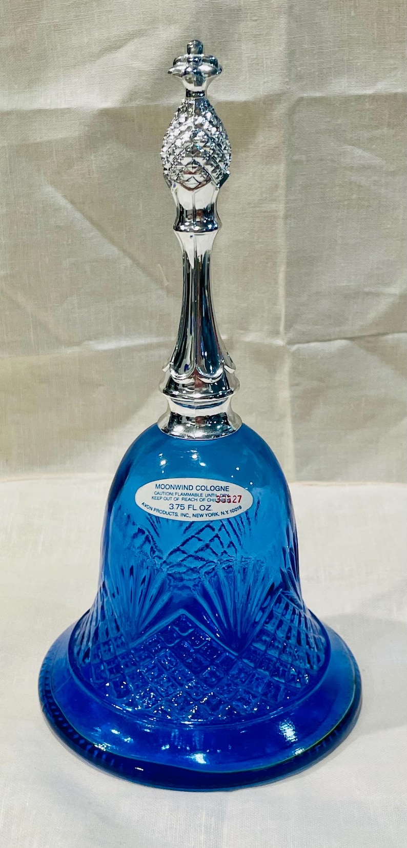 May include: A cobalt blue glass bell-shaped cologne bottle with a silver-colored metal top and stem. The bottle has a textured pattern and a label that reads "MOONWIND COLOGNE". The bottle is 3.75 fl oz.