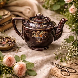 May include: A dark brown ceramic teapot with a floral design in gold and other colors. The teapot is on a beige cloth, surrounded by flowers, a bowl of tea leaves, and a pair of scissors. The teapot has a capacity of approximately 1.2 liters.