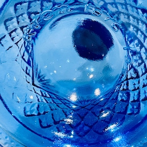 May include: Close-up of a vibrant blue glass object with a diamond pattern. The object has a circular shape with a textured surface and a central indentation. The glass reflects light, creating highlights and shadows.