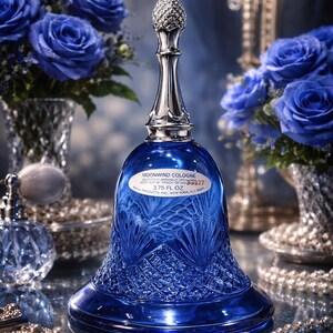May include: A cobalt blue cologne bottle with a silver pineapple-shaped stopper. The bottle is bell-shaped with a cut-glass design and the text "MOONWIND COLOGNE" and "3.75 FL OZ". Blue roses and a perfume bottle are in the background.