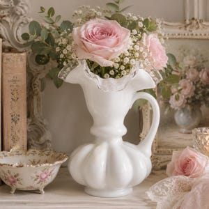 May include: A white, ruffled-edge pitcher holds pink roses and baby's breath. Beside it are vintage books, a floral bowl, a framed painting, and a lit candle, creating a romantic, vintage aesthetic.