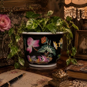 May include: A black ceramic planter featuring a colorful hand-painted design of pink lotus flowers, green lily pads, and small birds. The planter has a wide, flared rim and contains a trailing pothos plant with variegated green and white leaves.