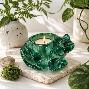 May include: A green glass frog-shaped candle holder with a lit white candle inside. The frog has detailed features and sits on a square stone. Decorative plants and small rocks are in the background.