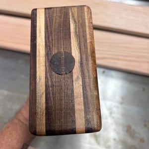 May include: A rectangular wooden mallet head featuring alternating vertical stripes of dark brown and light tan wood grain. A circular dark wood plug is centered on the face, displaying a visible seam running through the middle of the inlay.