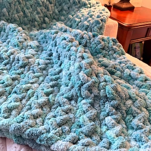 May include: A plush, hand-knitted blanket in shades of teal and light blue. The chunky knit creates a textured surface, perfect for cozy comfort. The blanket is folded over a bed, showcasing its soft, inviting appearance.