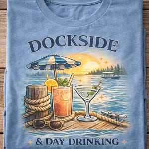 May include: Light blue t-shirt with a graphic of a dockside scene. The design includes a cocktail, martini, sunglasses, and the text "DOCKSIDE & DAY DRINKING". A parasol, pier, and boat are also featured.