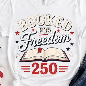 May include: White t-shirt with the text "BOOKED FOR Freedom" in a vintage style, featuring an open book graphic and the number 250. Red and blue stars and stripes are incorporated into the design. The shirt is paired with blue jeans.