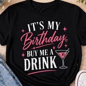 Op de afbeelding: Zwart T-shirt met de tekst "IT'S MY Birthday BUY ME A DRINK" in roze en wit. Het ontwerp bevat een martini glas afbeelding. Het shirt wordt gecombineerd met een blauwe jeans en witte sneakers.