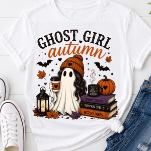May include: A white t-shirt featuring a graphic of a ghost wearing an orange beanie, holding a coffee cup, and standing next to a lantern, a pumpkin, and a stack of three books labeled Spooky Stories, Pumpkin Spells, and Witchy Vibes.