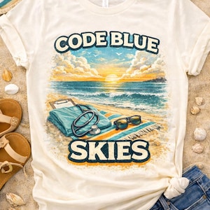 May include: Cream-colored t-shirt with a beach scene graphic. The design includes the words "CODE BLUE SKIES" above and below a depiction of a beach with medical scrubs, a stethoscope, and sunglasses. The shirt is paired with denim shorts and sandals.