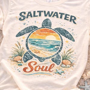 Puede incluir: Camiseta color crema con un diseño de tortuga y una escena de playa. El diseño incluye las palabras "Saltwater Soul" en verde azulado y naranja. Conchas marinas y estrellas de mar están esparcidas alrededor de la tortuga.