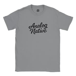 May include: A gray t-shirt with the words "Analog Native" in black script across the front. The shirt has short sleeves and a crew neck. The text "Mild Cliche Diem" is printed above the collar.