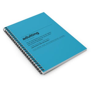 Op de afbeelding: Een felblauw spiraalnotitieboek met een woordenboekdefinitie van het woord adulting in zwarte tekst op de voorkant, inclusief de fonetische uitspraak en humoristische voorbeeldzinnen over het dagelijks leven.