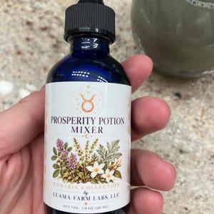 May include: A cobalt blue glass dropper bottle with a black cap and a white label. The label features the text Prosperity Potion Mixer, Lunaria Collection by Llama Farm Labs, LLC, and Net Vol. 2.0 oz (60 ml) with botanical illustrations.