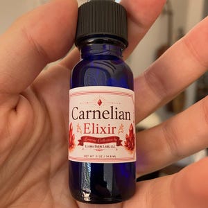 Könnte beinhalten: Eine kobaltblaue Glastropfflasche mit schwarzem Verschluss und weißem Etikett. Das Etikett zeigt den Text Carnelian Elixir, Lunaria Collection by LLAMA FARM LABS, LLC und NET WT .5 OZ / 14.8 ML sowie Abbildungen von roten Kristallen.