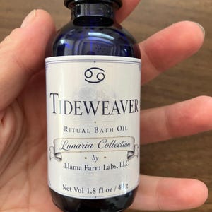 May include: A dark blue glass bottle with a black screw cap and a white label. The label features the text TIDEWEAVER RITUAL BATH OIL, Lunaria Collection by Llama Farm Labs, LLC, a Cancer zodiac symbol, and Net Vol 1.8 fl oz / 49 g.