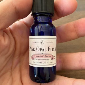 May include: A cobalt blue glass dropper bottle with a black cap and a white label. The label features the text Pink Opal Elixir, Lunaria Collection, by Llama Farm Labs, LLC, and Net 0.5 fl oz (15 mL) with a crescent moon and star graphic.