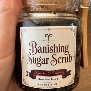 May include: A clear glass jar filled with dark brown sugar scrub, topped with a cork lid. The label reads Banishing Sugar Scrub, Lunaria Collection by Llama Farm Labs, LLC, with a net weight of 8 oz / 227 g printed at the bottom of the label.