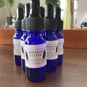 May include: A group of cobalt blue glass bottles with black droppers. Each bottle has a white label with the words "OBSIDIAN ELIXIR" and other text. The bottles are arranged on a wooden surface.